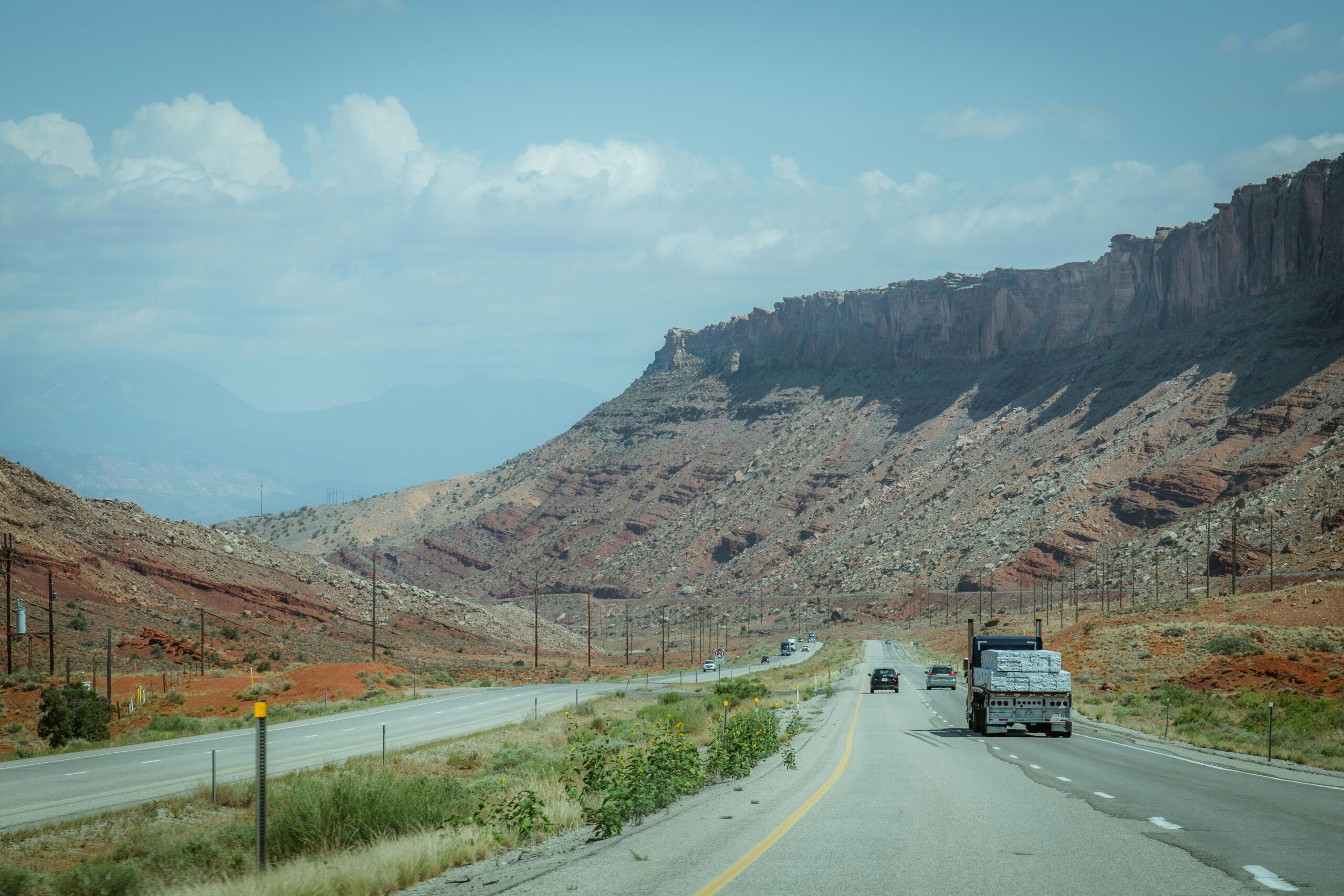 A truck drives on a desert highway surrounded by rugged mountains and clear skies.